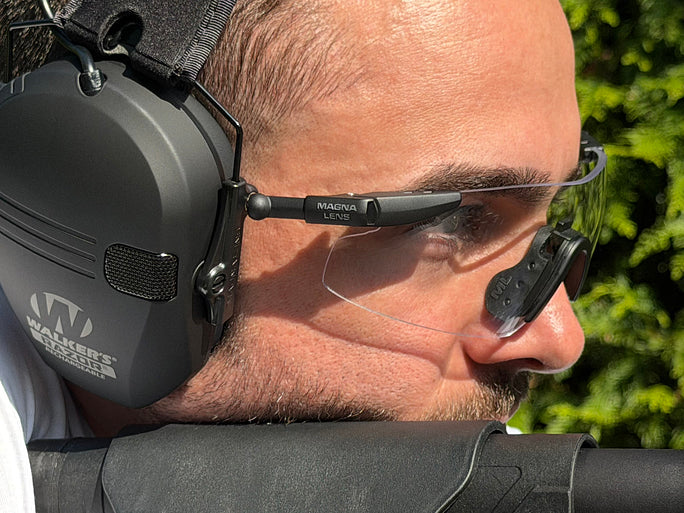 Close-up side profile of a man wearing Magnums clear shield glasses and a black Wagner hearing protector outdoors with green foliage in the background.